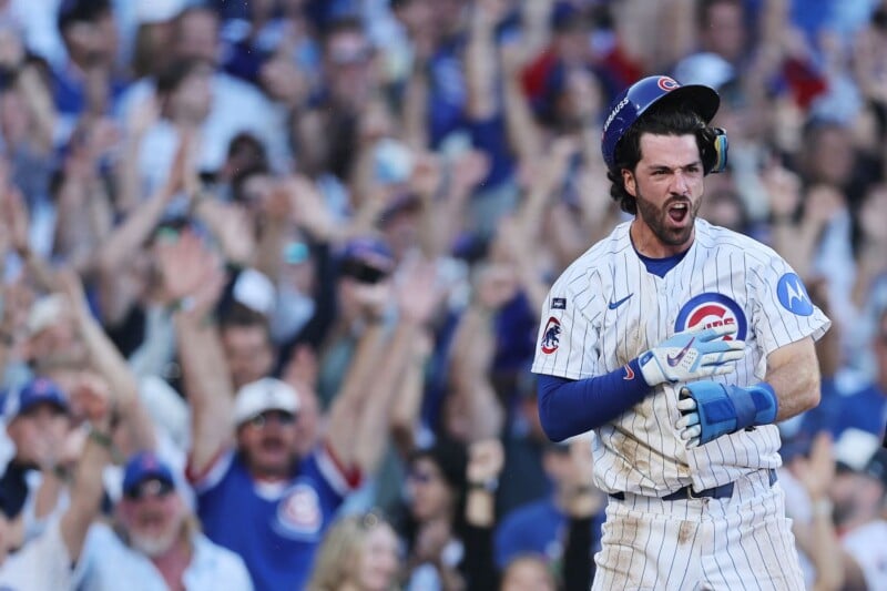 Chicago Cubs baseball player celebrates energetically on the field, pumping his fist and shouting, as a crowd of fans in the background cheer with raised arms at a stadium during a game.