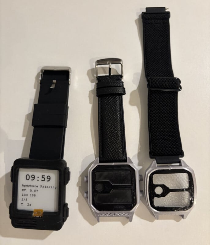 Tres relojes con pulsera negra se encuentran uno al lado del otro sobre una superficie blanca. El reloj de la izquierda tiene una pantalla digital con ajustes de cámara y los otros dos tienen esferas en blanco o estilizadas con patrones geométricos.