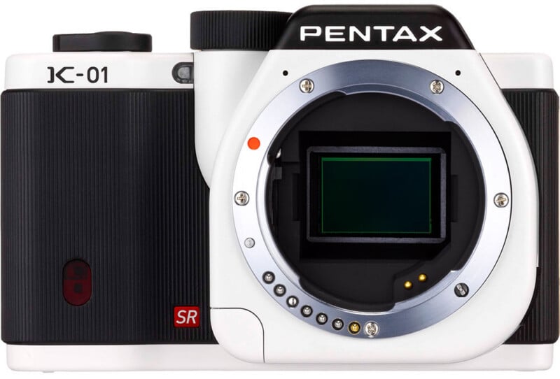 A white and black Pentax K-01 digital camera body, shown from the front with no lens attached, revealing the sensor and lens mount.