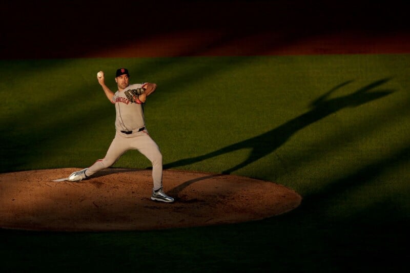 A baseball pitcher in a gray uniform throws a pitch on a mound, his shadow dramatically cast on the grass by the setting sun.