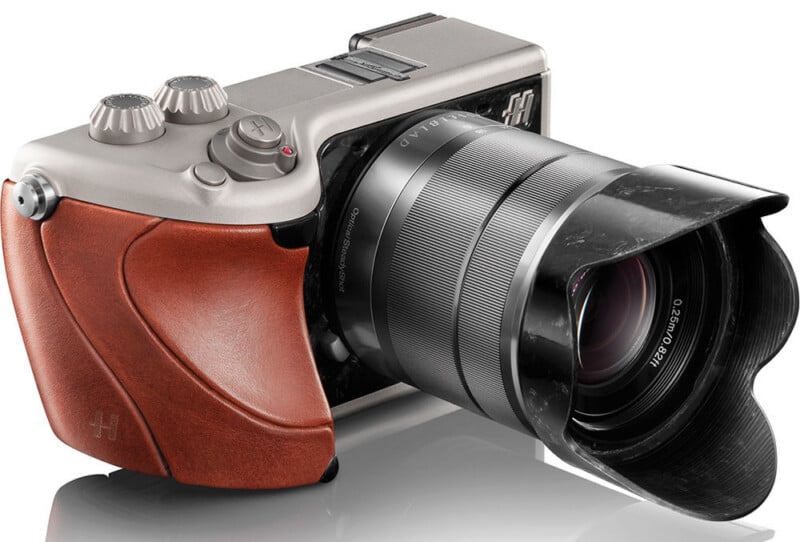 A modern digital camera with a silver body, brown leather grip, and a large detachable lens with a lens hood, placed on a reflective white surface.