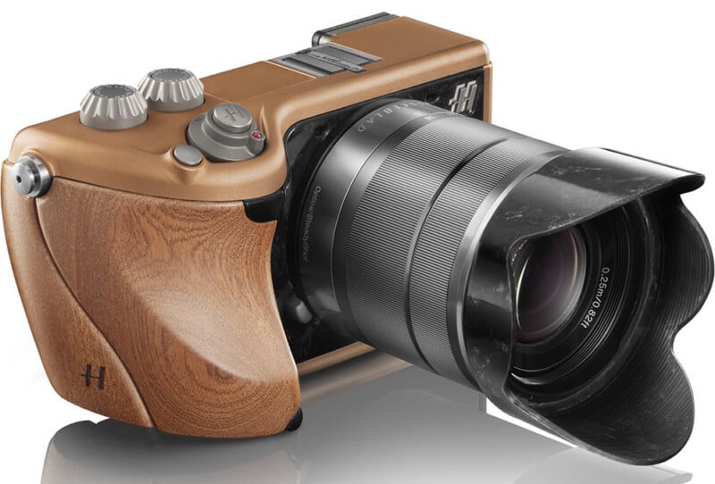 A stylish camera with a brown wooden grip, two silver control dials, and a large black lens with a petal-shaped hood, set against a white background with a soft reflection below.