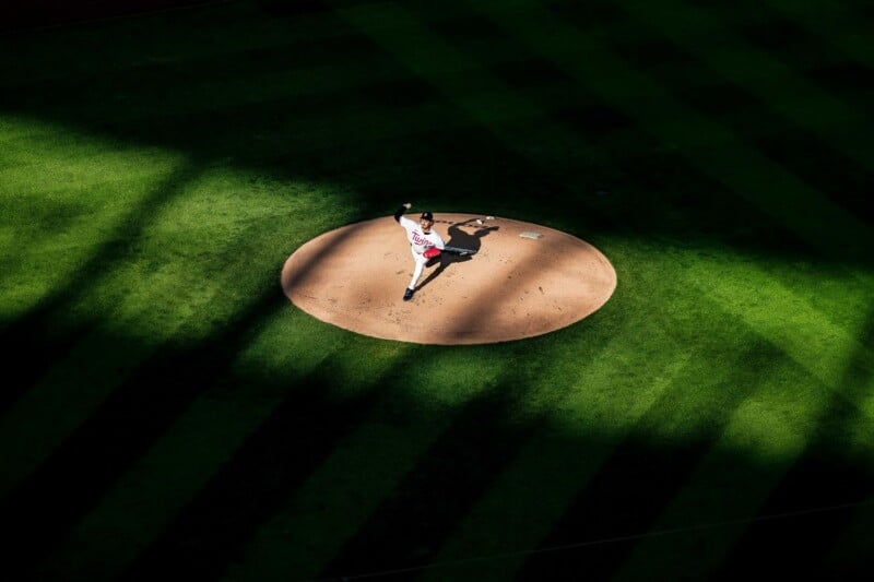 A baseball pitcher in a white uniform throws from the mound, spotlighted by sunlight, surrounded by dark shadows and striped grass on the field.