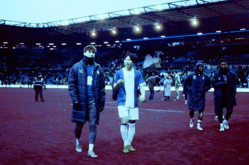 Two soccer players in jackets and uniforms walk across a maroon-colored field in a stadium, with several other players and empty blue seats visible in the background under bright lights.