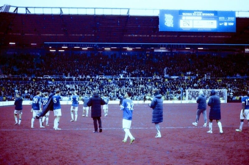 Football players in blue jerseys walk across a red-tinted field in a stadium filled with spectators. Some staff members are present, and a scoreboard is visible in the background. The atmosphere appears energetic.