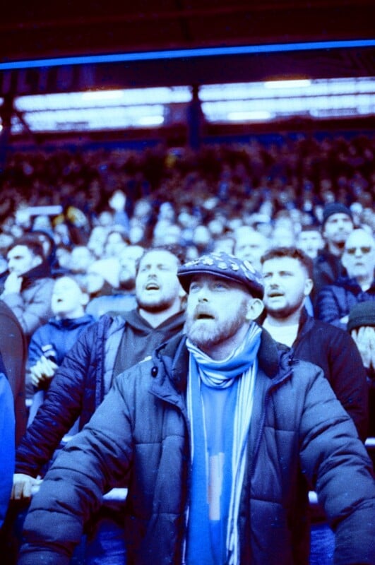 A bearded man in a scarf and cap stands in a crowded stadium, looking intently ahead. Behind him, a lively crowd of fans appears focused on the event, with blue lighting casting a cool tone over the scene.