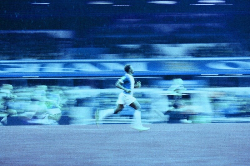 A motion-blurred image shows a soccer player in blue sprinting across the field, with other players and a stadium full of spectators in the background under blue-tinted lighting.