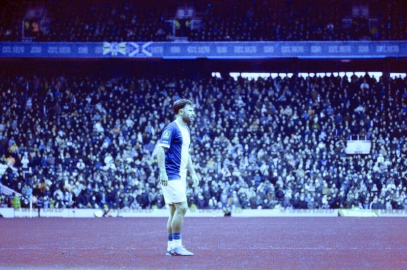 A soccer player in a blue and white uniform stands on a red-tinted field, with a large crowd of spectators filling the stadium in the background.