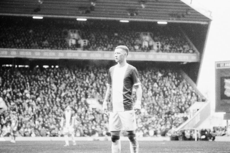 A black and white photo of a soccer player standing on the field during a match, with a blurred crowd filling the stadium in the background.