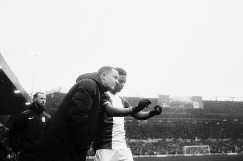 A black-and-white photo shows two soccer players on a stadium sideline, one in a tracksuit and the other in a team jersey, talking and gesturing toward the field while a crowd fills the stands in the background.