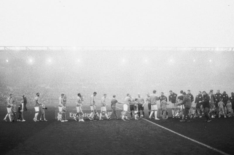 Two teams of soccer players line up and shake hands on a foggy field before a match, with spectators in the stands barely visible through the mist in the background.