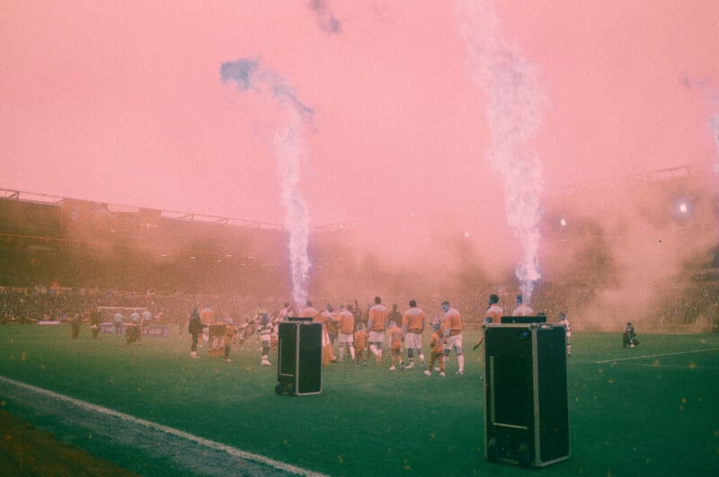 Soccer players walk onto a field amid smoke and blue flames, with a large crowd in the background and a pink-orange tint over the scene, creating a dramatic atmosphere.
