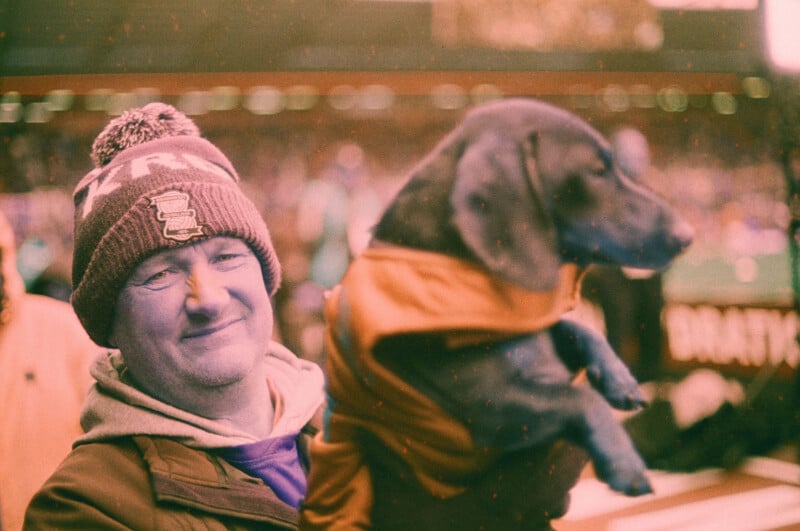 A man in a winter hat smiles while holding a small dog dressed in a coat. They are outdoors, with a blurred stadium crowd in the background. The image has a warm, vintage tone.