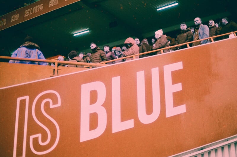 A group of people in winter clothing stand on a stadium balcony above a large red sign that reads "IS BLUE" in bold white letters.