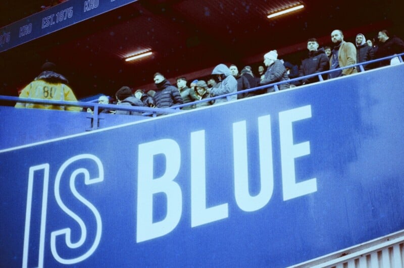 A group of people dressed in winter clothing stand on a stadium balcony above a large blue sign that reads "IS BLUE." A security guard in a yellow vest stands nearby.