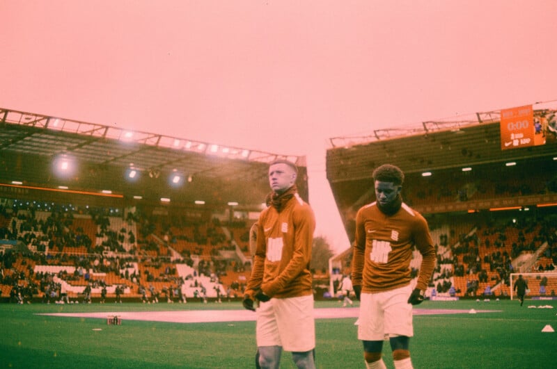 Two soccer players in warm-up attire stand on a field in a stadium with many empty seats. The image has a reddish-orange tint, and an electronic scoreboard is visible in the background.