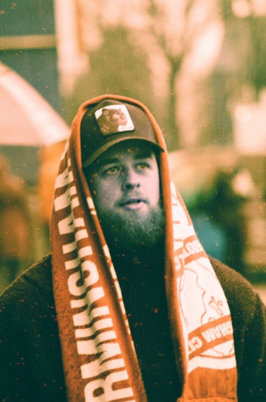 A bearded man wearing a cap stands outdoors with a red and white scarf draped over his head and shoulders. The image has a warm, vintage filter and blurred background with indistinct people and trees.