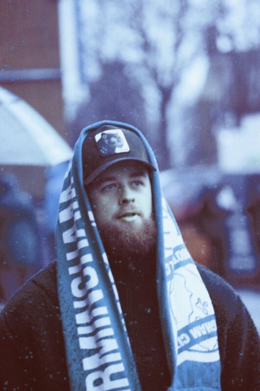 A bearded man wearing a dark cap and a blue-and-white scarf stands outdoors on a snowy day. The scarf is draped over his head, and people and trees are blurred in the background.