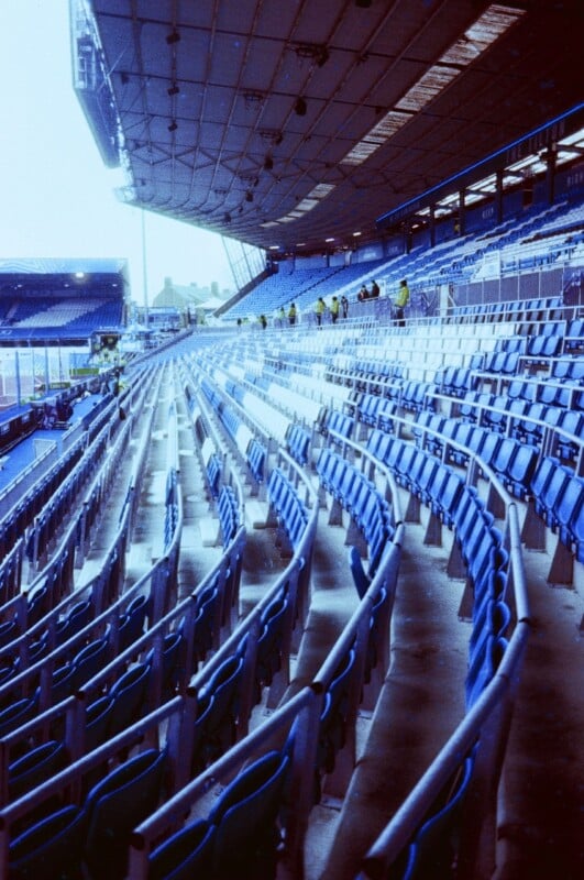 Rows of empty blue stadium seats curve under a large, partially covered roof, with a few people visible in the distance near the top of the stands. The setting appears quiet and awaiting an event.