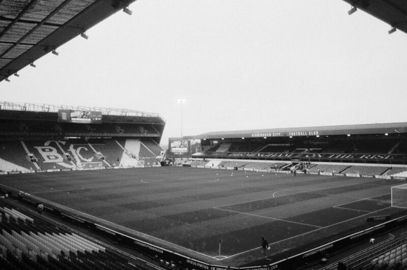 Wide-angle view of an empty football stadium in black and white, with rows of seats surrounding a well-maintained pitch and large stands labeled "BCFC" and "Birmingham City Football Club.