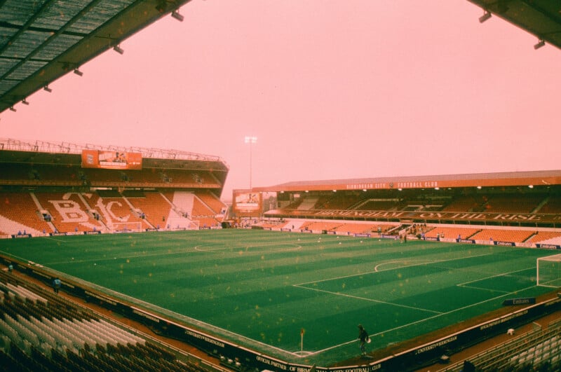 A large, mostly empty football stadium with red seating and green field under a pink-tinted sky. The stands read “BCFC,” and a scoreboard displays information on one side. The stadium lights are on.