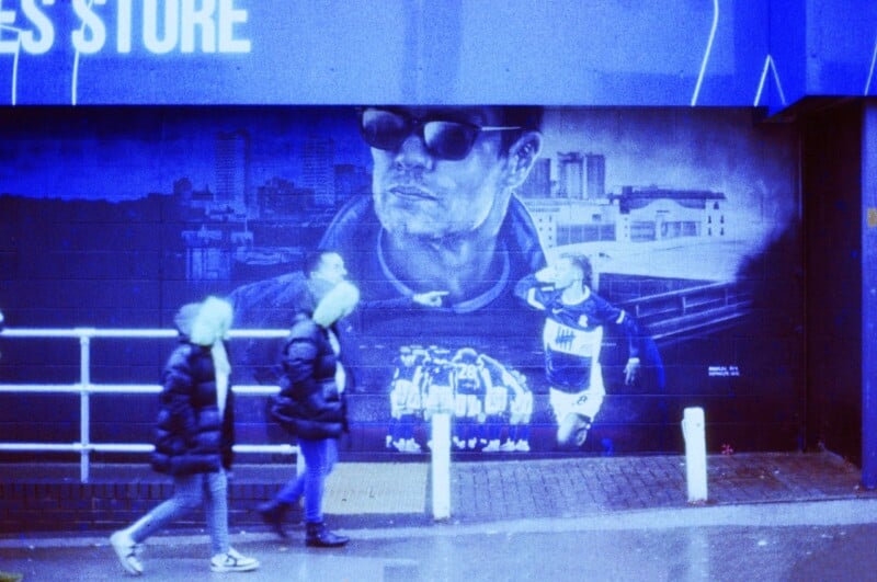 Two people in winter coats walk past a blue-toned mural depicting a man with sunglasses, a group of soccer players, and a cityscape background. The mural is painted on a building’s wall.