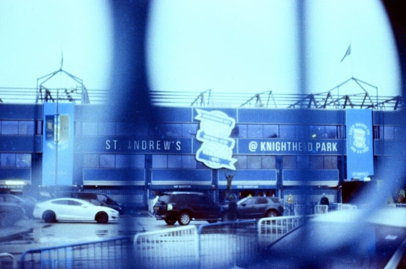 A blue stadium building with "St. Andrew's @ Knighthead Park" on the front, several parked and moving cars in the foreground, and blurry blue bars partially blocking the view.