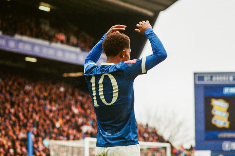 A soccer player wearing a blue jersey with the number 10 raises his arms in front of a cheering stadium crowd during a daytime match.