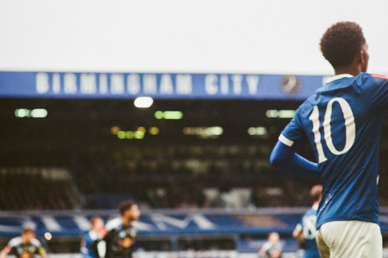 A soccer player wearing a blue jersey with the number 10 stands on the field, with the blurred Birmingham City stadium and other players in the background.