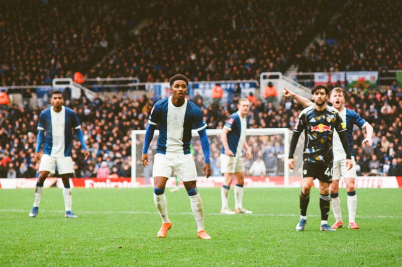 Several soccer players from two opposing teams stand alert on the field during a match, with a packed stadium and spectators visible in the background.