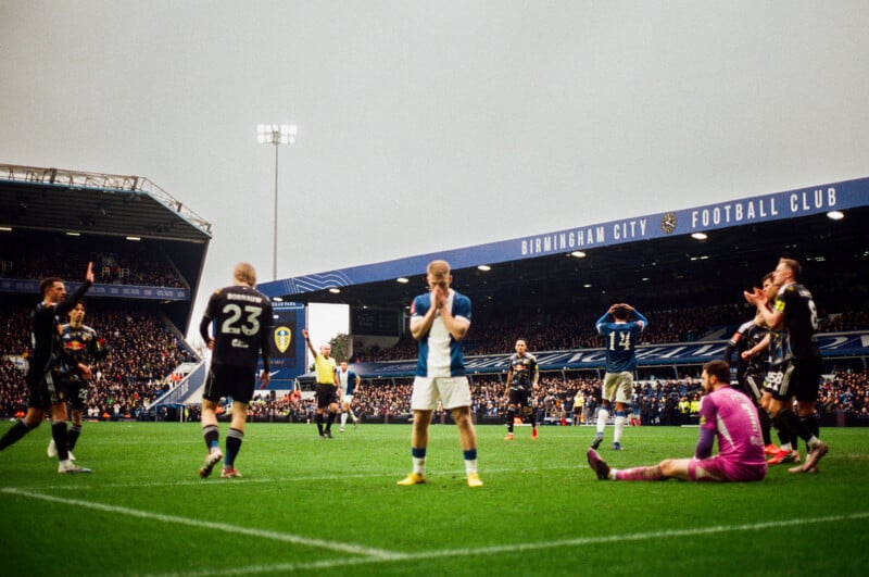 A football player in blue and white stands with his hands covering his face in disappointment, while others react around him on the field at Birmingham City Football Club’s stadium during a match.