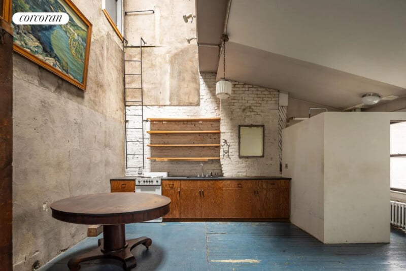 A rustic kitchen with a round wooden table, blue-painted wooden floor, open shelves, a small stove, brick and plaster walls, and a painting on the wall. Natural light enters through high windows.