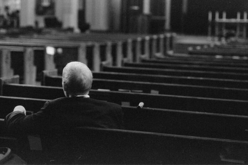 A black-and-white photo shows an older man with light hair sitting alone in a mostly empty row of wooden pews, facing forward in a large, dimly lit room that appears to be a church or auditorium.