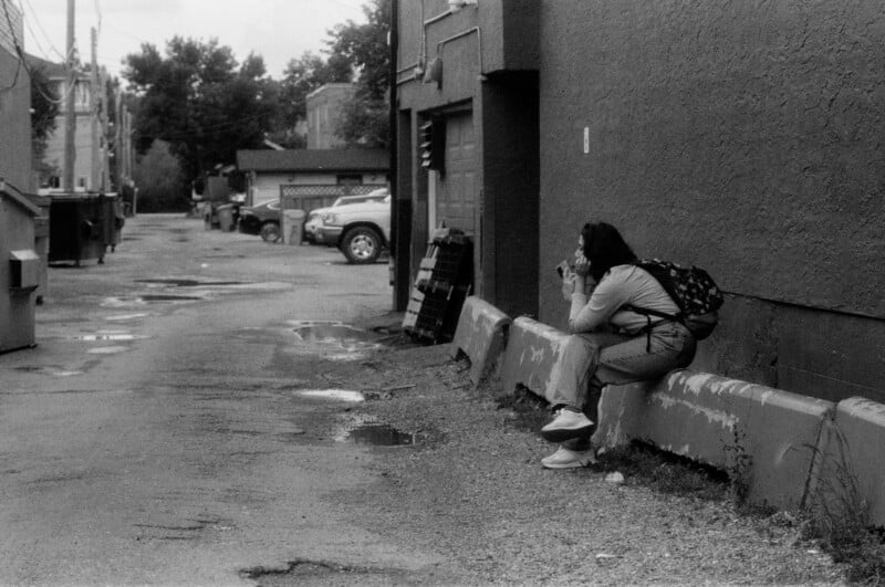 A person with a backpack sits alone on a concrete barrier in a deserted alley, eating or drinking, with parked cars and puddles visible in the background. The scene is in black and white.