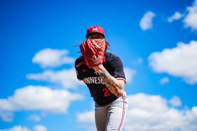 A baseball pitcher in a Minnesota uniform prepares to throw a pitch, holding a red glove up to his face against a bright blue sky with scattered clouds.