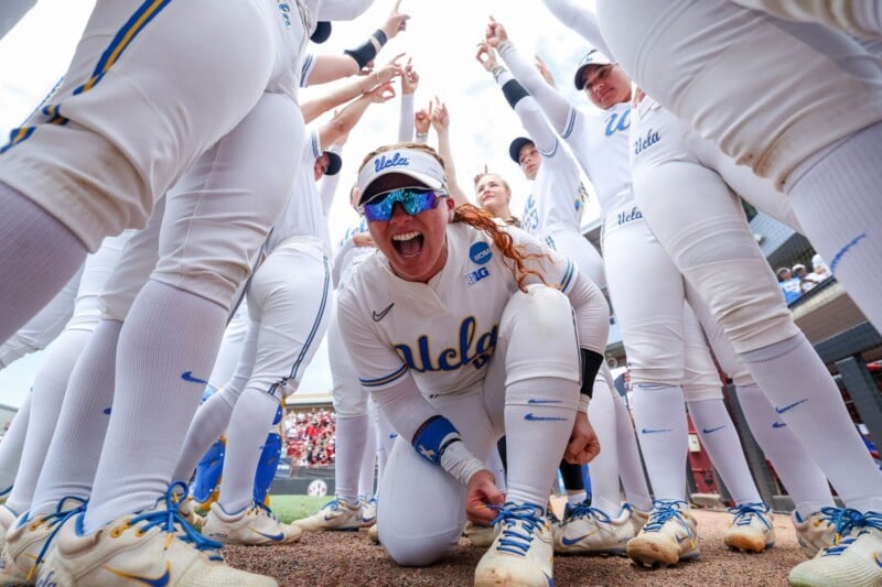 A group of UCLA softball players in white uniforms stand in a huddle, smiling and cheering, with one player crouching in the center, pointing and celebrating enthusiastically.