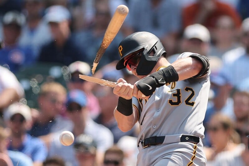 A baseball player in a gray uniform swings and shatters his bat, with the ball and broken bat pieces flying through the air during a game, as a blurred crowd watches in the background.