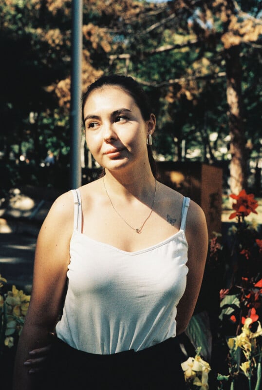 A young woman with dark hair tied back stands outdoors among flowers, wearing a white tank top and necklace, looking slightly to the side with a thoughtful expression. Sunlight casts soft shadows on her face and shoulders.