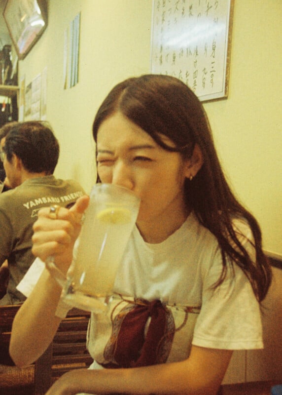 A young woman with long dark hair is sitting indoors, squinting one eye as she drinks a glass of lemonade with a slice of lemon. Japanese writing is visible on a sign behind her.