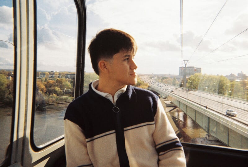 A young person with short hair sits in a cable car, looking out the window. Behind them, a river, trees, and a road with cars are visible under a partly cloudy sky.