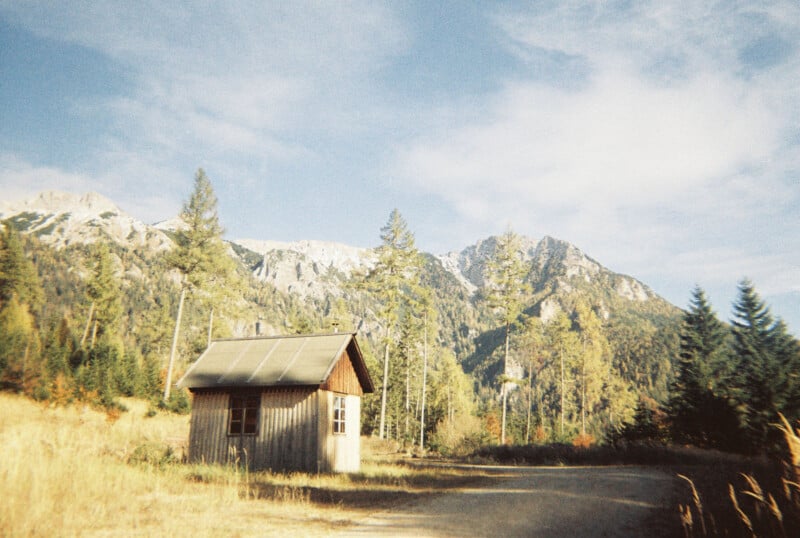 A small wooden cabin sits in a sunlit clearing, surrounded by tall trees and mountains under a partly cloudy sky. A dirt path curves beside the cabin, and the grass is golden, suggesting late summer or early autumn.