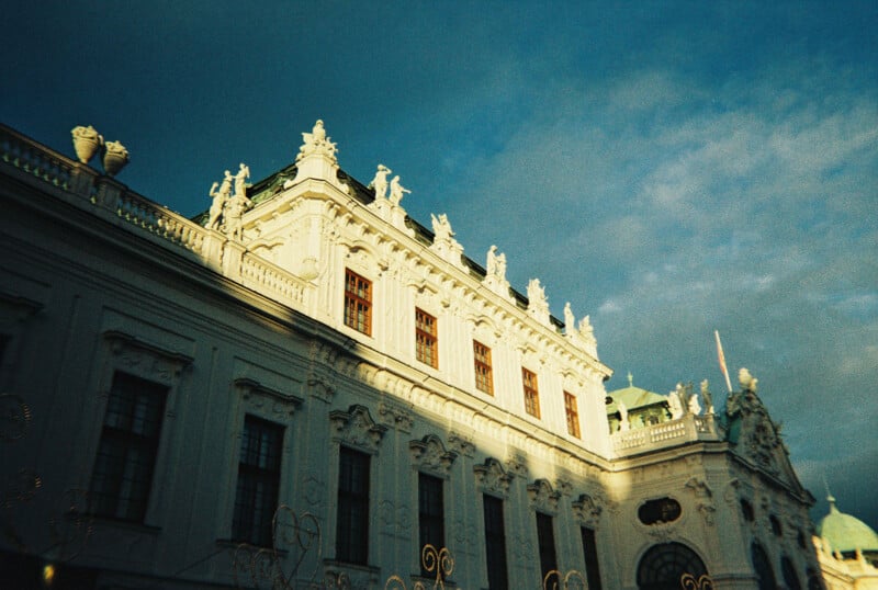 An ornate historic building with statues along its roofline is lit by sunlight, with dramatic shadows on the facade and a partly cloudy blue sky overhead.