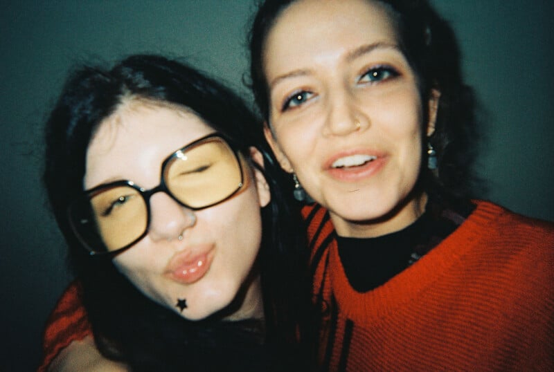 Two young women pose closely together. One wears large tinted glasses, puckers her lips, winks, and has a star tattoo near her mouth. The other smiles, wearing a red sweater and hoop earrings. Both appear happy and relaxed.