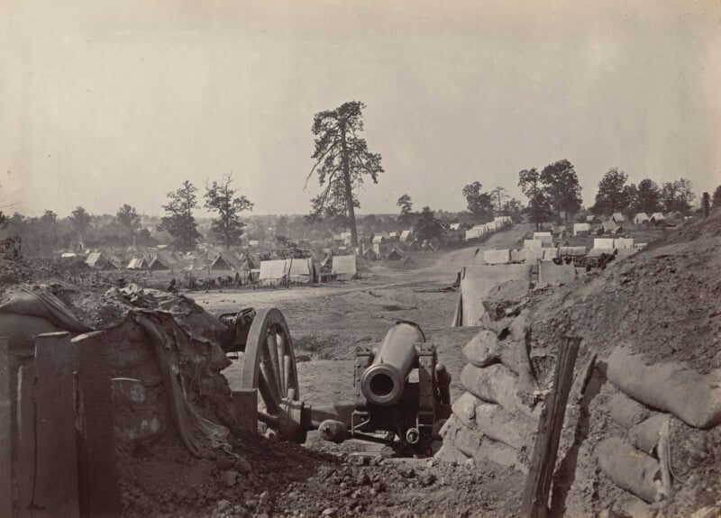 Una fotografía en blanco y negro muestra un campamento militar de la época de la Guerra Civil con tiendas de campaña esparcidas por el campo. En primer plano, detrás de sacos de arena y movimientos de tierra, hay un cañón. El camping a lo lejos está rodeado de árboles.