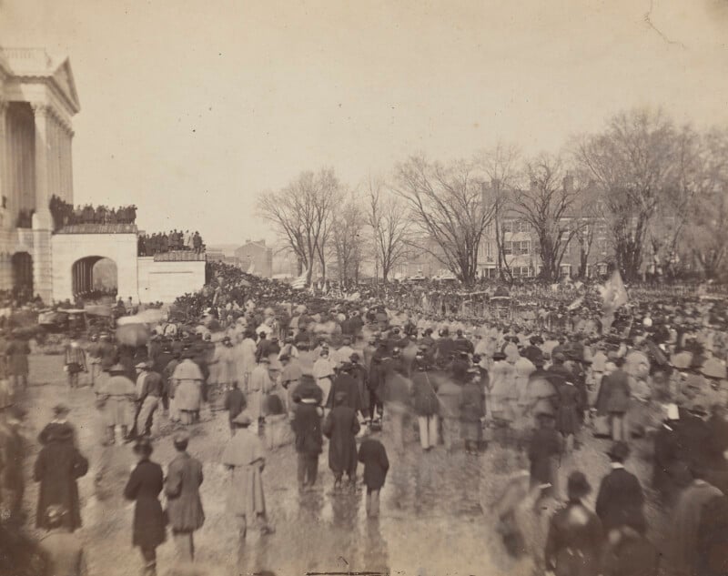 En un día de invierno, una gran multitud de personas, muchas de ellas con abrigos y sombreros, se reunieron frente a un gran edificio con columnas. Al fondo se ven árboles desnudos y otros edificios.