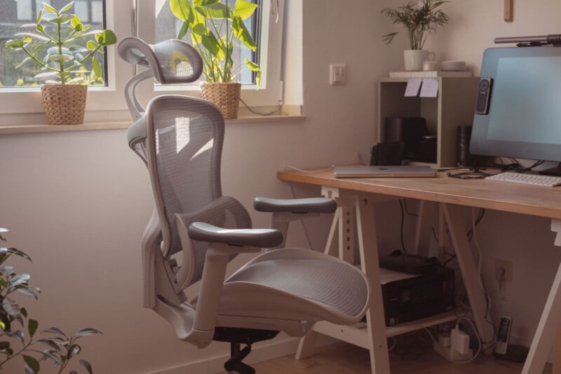 A modern ergonomic office chair sits in front of a wooden desk with a computer in a bright room. Sunlight streams through the window, and potted plants are on the windowsill and desk, creating a cozy workspace.