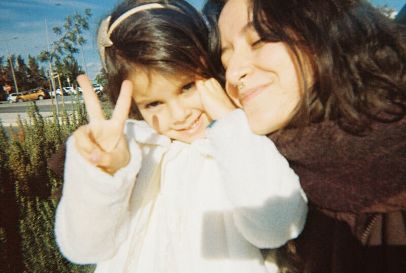 A young woman and a little girl smile closely together outdoors. The girl, wearing a white coat, holds up a peace sign, while the woman, wearing a scarf, has her eyes closed and is smiling.