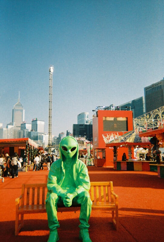 A person in a bright green alien costume sits on a wooden bench in an outdoor fairground, surrounded by colorful booths and city skyscrapers under a clear blue sky.