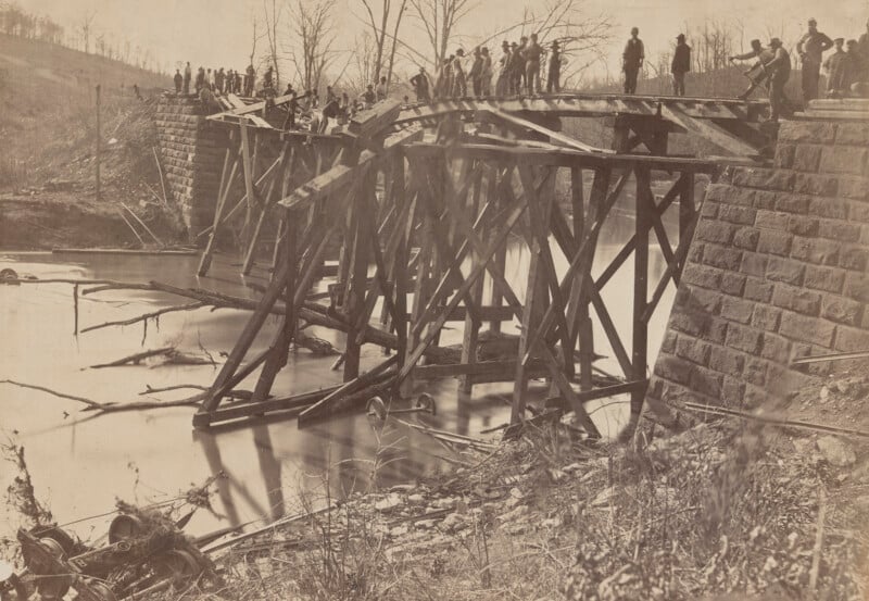 Un grupo de personas se encontraba sobre y alrededor de un puente de madera dañado sobre el río con secciones derrumbadas y escombros en el agua, rodeado de árboles desnudos y un terraplén rocoso.