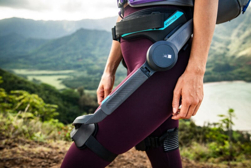 A person stands outdoors wearing a high-tech exoskeleton device on their hip and thigh, overlooking a lush mountainous landscape and a body of water in the distance.